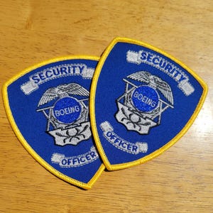 May include: Two blue and yellow embroidered patches with the words "SECURITY OFFICER" and the Boeing logo. The patches are shaped like a shield.
