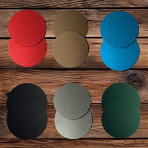 May include: A collection of twelve circular coasters arranged in two rows. The coasters feature a solid, textured finish in various colors including red, brown, blue, black, gray, and dark green. Each coaster is a simple, flat, round disc shape.