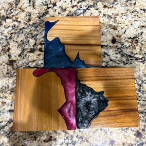 May include: Two wooden coasters with a natural wood grain. One coaster has a dark blue resin design, while the other has a magenta and black resin design. The coasters are square and rectangular.