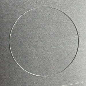 May include: A clear, circular lens or glass, possibly for eyeglasses or a similar optical device, is centered against a textured, metallic gray surface. The thin, transparent circle is the primary focus, with the background providing a subtle contrast.