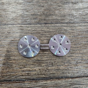 May include: Two silver metal discs with eight holes each, connected by two silver metal rods. The discs have a shiny, reflective surface.