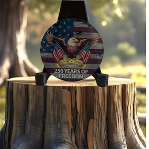 May include: A circular decorative plaque featuring a bald eagle with spread wings, two American flags, and the text 1776-2026 250 YEARS OF FREEDOM. The design includes a distressed American flag background and is displayed on a black stand.