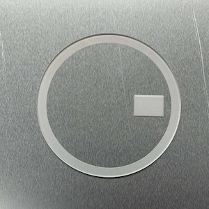 May include: A clear, circular object with a rectangular shape inside. The object is set against a textured, metallic background. The circular shape has a thin, transparent border.