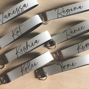 May include: Seven white leather keychains with black lettering. The keychains are personalized with the names Vanessa, Kat, Keshia, Koley, Toni, Regina, and Renee.