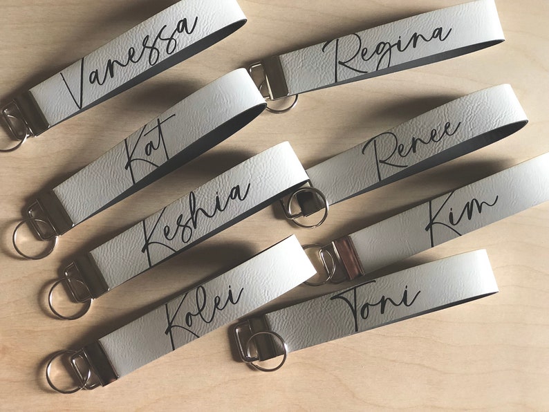 May include: Seven white leather keychains with black lettering. The keychains are personalized with the names Vanessa, Kat, Keshia, Koley, Toni, Regina, and Renee.