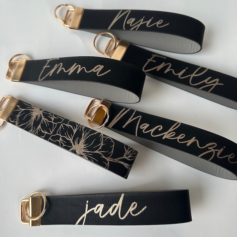 May include: Six black leather keychains with gold metal rings and gold lettering. The keychains are personalized with the names "Emma", "Emily", "Mackenzie", "Jade", "Najie", and a floral design.