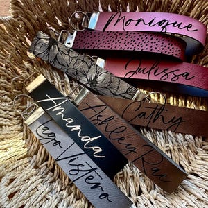 May include: A collection of six leather keychains with different colors and names printed on them. The names are "Monique", "Julissa", "Cathy", "Ashley Rae", "Amanda", and "Lego Visiera".
