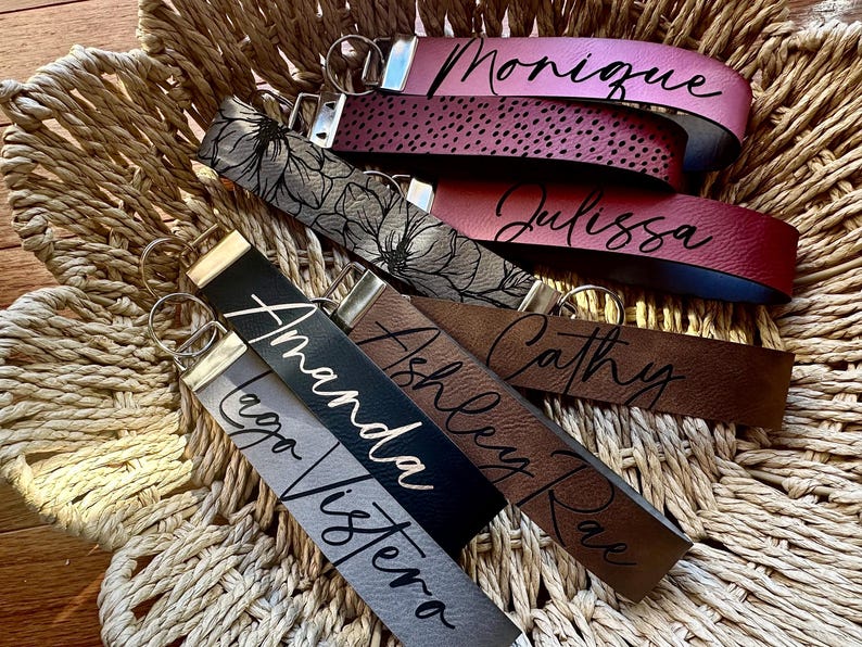 May include: A collection of six leather keychains with different colors and names printed on them. The names are "Monique", "Julissa", "Cathy", "Ashley Rae", "Amanda", and "Lego Visiera".