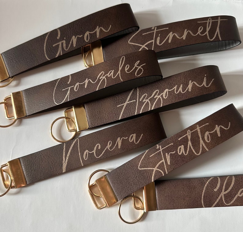 May include: Six brown leather keychains with gold key rings. Each keychain has a different name engraved in gold lettering: Girion, Stinnett, Gonzales, Azzouni, Nocera, Stratton.