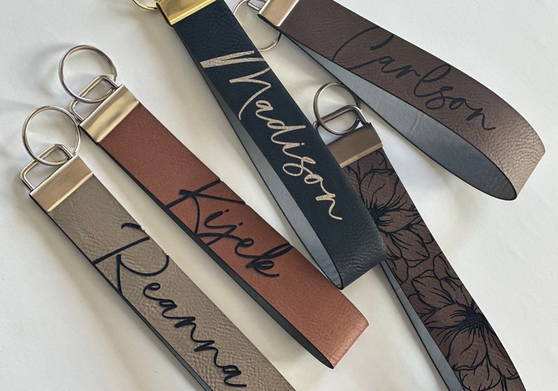 May include: Four personalized leather keychains with metal rings. The keychains are brown, black, gray, and brown with a floral pattern. The names "Reama", "Kiziek", "Madison", and "Carlson" are engraved on the keychains.