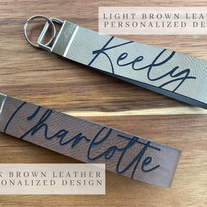 May include: Two leather keychains with personalized names. The top keychain is light brown leather with the name "Keely" written in black cursive. The bottom keychain is dark brown leather with the name "Charlotte" written in black cursive.