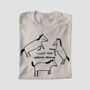 Op de afbeelding: Een lichtbeige T-shirt met ronde hals en een zwarte lijntekening van drie paarden in verschillende houdingen. De tekst "I CAN'T STOP HORSING AROUND" is in het zwart in het midden van het T-shirt gedrukt, tussen de drie afgebeelde paarden.