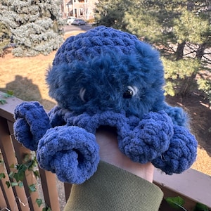 Puede incluir: Un pulpo de crochet azul con pelusa suave en la cabeza. El pulpo tiene ojos grandes y blancos y está siendo sostenido por una mano.