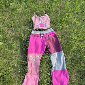 May include: A dusty pink spaghetti strap crop top featuring a central star appliqué and a pair of color-blocked sweatpants. The pants include panels of magenta, dark maroon, and grey, with a drawstring waist and a small graphic print on the leg.