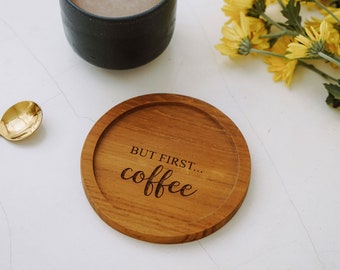 May include: A wooden coaster with the text "BUT FIRST... coffee" engraved on it. The coaster is brown and has a natural wood finish. A cup of coffee and a gold spoon are also in the image.