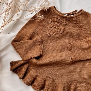 Può includere: Un maglione marrone lavorato a maglia con maniche lunghe e orlo a balze. Il maglione presenta un motivo floreale sul petto e un dettaglio a fiocco sulla schiena. Il capo è realizzato in un filato morbido e strutturato.