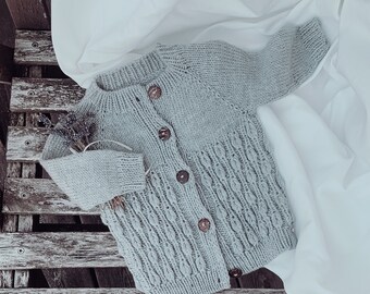 May include: A light blue knitted cardigan with wooden buttons. The cardigan is laid out on a wooden surface with a white fabric background.