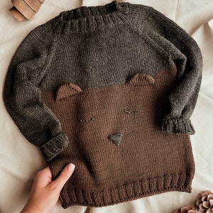Può includere: Un maglione di lana marrone e verde con un disegno di un viso di orso. Il maglione ha un collo rotondo e maniche lunghe.