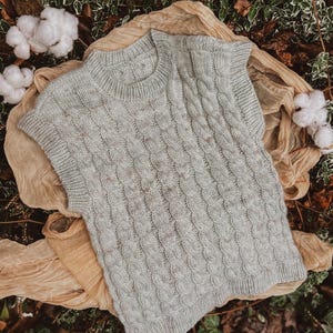 Könnte beinhalten: Hellgraue Strickweste mit Zopfmuster und Rundhalsausschnitt. Das ärmellose Oberteil hat ein strukturiertes Muster und wird auf einem beigen Stoff mit Baumwollakzenten präsentiert. Die Weste hat eine weiche, neutrale Farbe.