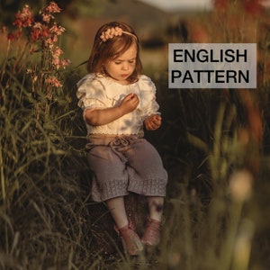 May include: A young girl wearing a white blouse with a ruffled collar and a light purple knitted skirt sits on a rock in a field of tall grass. She is wearing a pink flower headband and has a thoughtful expression on her face. The text "ENGLISH PATTERN" is visible in the upper right corner of the image.