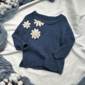 Könnte beinhalten: Ein blauer Strickpullover mit weißen gehäkelten Blumenverzierungen.