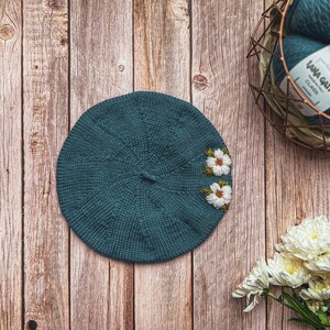 May include: A teal blue knitted beret with white embroidered flowers. The beret is lying on a wooden surface.