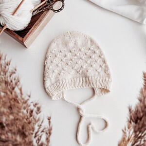 Könnte beinhalten: Eine weiße gestrickte Baby-Mütze mit Rautenmuster und einem Band am unteren Rand.