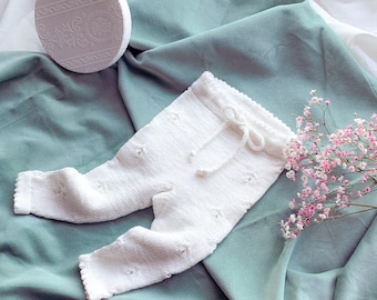 Op de afbeelding: Witte gebreide babybroek met een trekkoord in de taille en een delicaat bloemenpatroon. De broek ligt op een groene stoffen achtergrond.