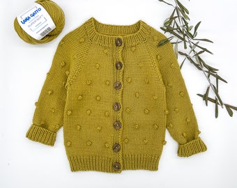 May include: A mustard yellow knitted cardigan with a button closure and a textured bobble stitch. The cardigan has long sleeves and a round neckline.