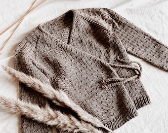 May include: A brown knitted cardigan with a wrap style and a tie closure. The cardigan is made with a textured stitch and has a subtle speckled pattern. Two skeins of brown yarn are visible in the background.