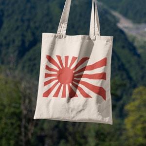 May include: A white canvas tote bag with a red rising sun design. The bag has two handles and is being held by a person's hand.
