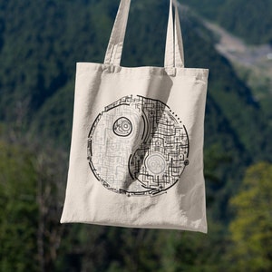 May include: A beige tote bag featuring a black and grey yin yang design. The design incorporates a circuit board pattern. The bag is held up against a backdrop of mountains and a blue sky.