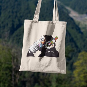May include: A white canvas tote bag with a black and white illustration of a monkey wearing headphones. The monkey is holding a microphone in one hand and has a colorful design on its face.