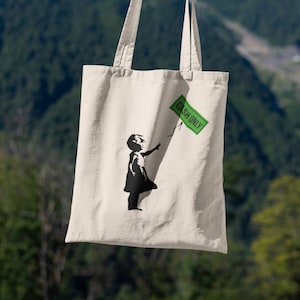 Peut inclure: Sac fourre-tout crème avec une silhouette noire d'une fille tendant la main vers un signe vert "CASH ONLY". Le sac est tenu devant un fond de montagnes et de ciel bleu.