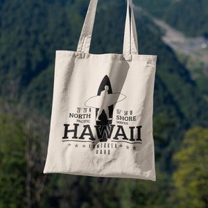 May include: A white canvas tote bag with black text and a silhouette of a surfer. The text reads "21 28 N NORTH PACIFIC 157 58 W SHORE WAVES HAWAII *LANIAKEA* OAHU".