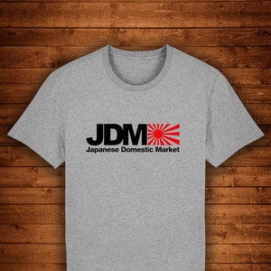 Puede incluir: Una camiseta gris jaspeado con el texto "JDM" en negro, con un diseño de sol naciente rojo y blanco. Debajo, la frase "Japanese Domestic Market" en negro. La camiseta se muestra sobre un fondo de madera.