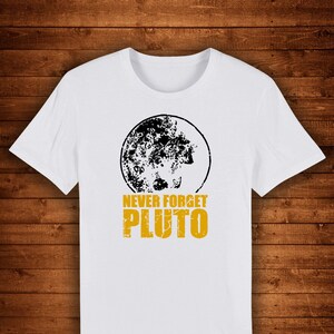 May include: White t-shirt featuring a black and white graphic of a planet with the text "NEVER FORGET PLUTO" in yellow. The shirt has a crew neck and short sleeves. The design is centered on the front of the shirt.