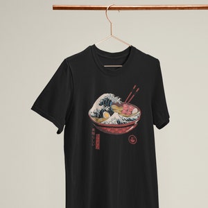 Peut inclure: T-shirt noir avec un graphique d'un bol de ramen avec une vague qui s'écrase dessus. Le texte "美味しい" est imprimé sur le t-shirt.
