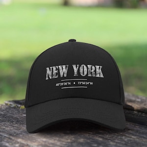 Può includere: Cappellino da baseball nero con la scritta "NEW YORK" in un carattere strutturato e usurato. Sotto, le coordinate 40°39'36"N 73°56'24"W. Il cappellino è su una superficie di legno.