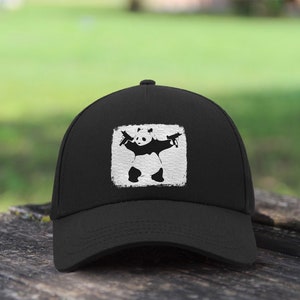 Può includere: Cappellino da baseball nero con una grafica bianca di un panda che tiene due pistole. La grafica è in un quadrato bianco con un aspetto usurato.