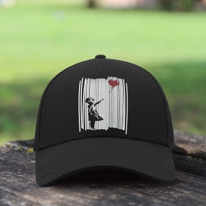Könnte beinhalten: Schwarze Baseballkappe mit einem weißen und schwarzen Grafikdruck eines Mädchens, das einen roten Herzluftballon hält. Die Grafik ist von weißen vertikalen Linien umgeben.