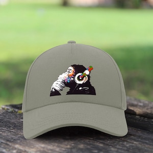 Può includere: Un berretto da baseball grigio con una grafica colorata di una scimmia che indossa le cuffie e pensa. La scimmia è circondata da schizzi di vernice colorati.