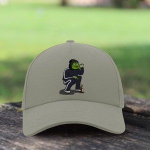 Puede incluir: Una gorra de béisbol beige con una imagen bordada de un personaje de dibujos animados verde que lleva una sudadera con capucha y pantalones negros, sosteniendo una botella y un cigarrillo.