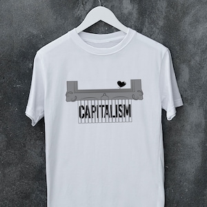 Könnte beinhalten: Weißes T-Shirt mit einem grafischen Design, das einen Aktenvernichter zeigt, der das Wort "KAPITALISMUS" schreddert. Ein kleines schwarzes Herz befindet sich über dem Schredder. Das T-Shirt hängt an einem weißen Kleiderbügel vor grauem Hintergrund.