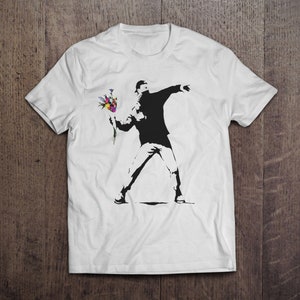 May include: White t-shirt featuring a black and white graphic print of a person throwing a bouquet of flowers. The flowers are colourful and include red, pink, yellow, blue, and purple.