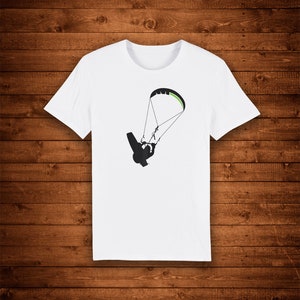 Puede incluir: Camiseta blanca con una silueta negra de parapente y un diseño de ala de parapente verde y negra. El diseño está centrado en el pecho. La camiseta tiene cuello redondo y mangas cortas.