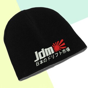 Op de afbeelding: Zwarte beanie met grijze rand, met de witte tekst "JDM" en Japanse karakters, plus een rood zonnestraalontwerp. De beanie is gemaakt van gebreid materiaal.