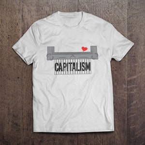Könnte beinhalten: Weißes T-Shirt mit einem grafischen Design. Das Design zeigt das Wort "CAPITALISM" in fetter, schwarzer Schrift, mit einem roten Herzen und einer grauen Illustration darüber. Das T-Shirt wird auf einer Holzoberfläche präsentiert.