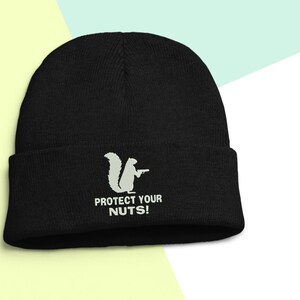 May include: Black beanie with a white squirrel graphic holding a gun and the text "PROTECT YOUR NUTS!". The beanie is made of a soft, knitted material.