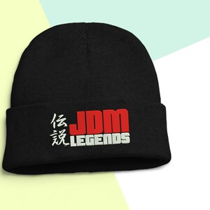 May include: Black beanie with the text "JDM LEGENDS" in red and white. The beanie has a folded cuff and a soft, knitted texture. The Japanese characters are embroidered in white.